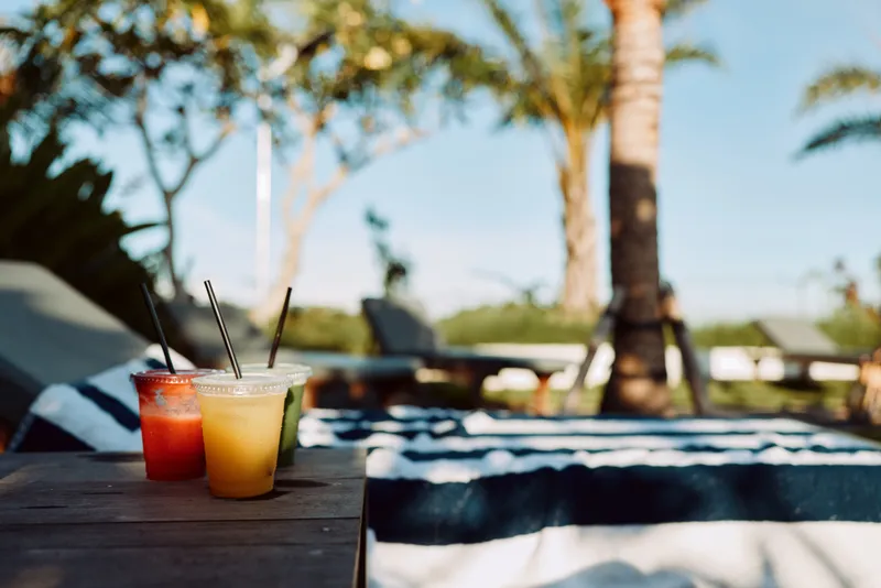 Two refreshing summer drinks with straws stand on a wooden table near striped towels by the pool under palm trees on a sunny day, creating a relaxed lifestyle atmosphere.