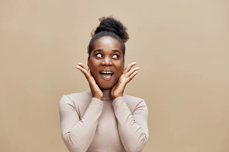 Happy young African woman expressing joy with hands on her cheeks, against a warm beige background, perfect for wellness and positivity concepts