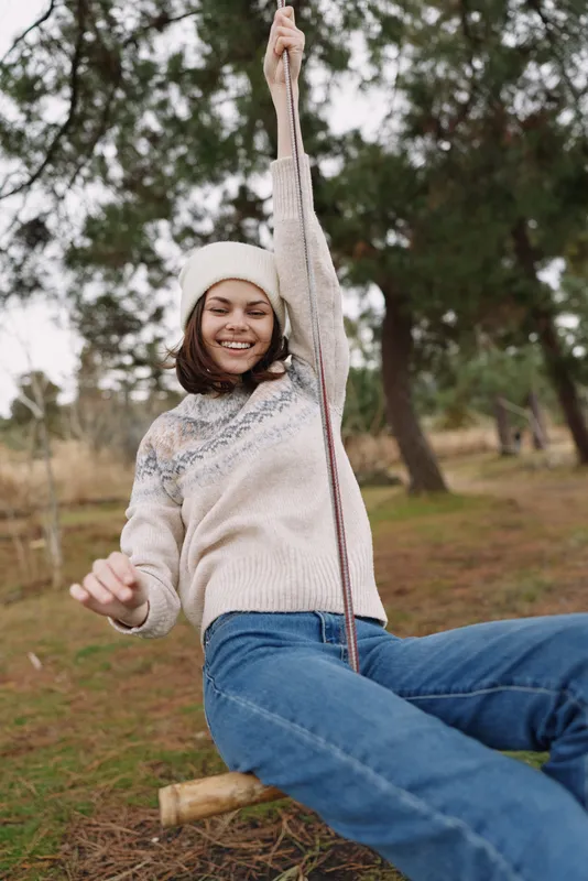 Girl on a wooden swing outdoors, smiling and carefree in a knit sweater and hat, surrounded by trees and autumn air, capturing playful joy and natural energy