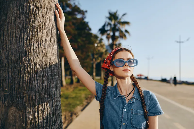 Lifestyle woman in modern boho-western denim street style posing confidently by a palm tree with film color effect and oversized sunglasses on sunny day.