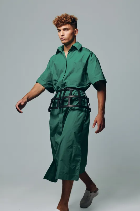 Young man walking in green oversized shirt dress with black belt accessory and casual shoes on gray background. Fashion model with curly hair posing in studio for style and clothing.
