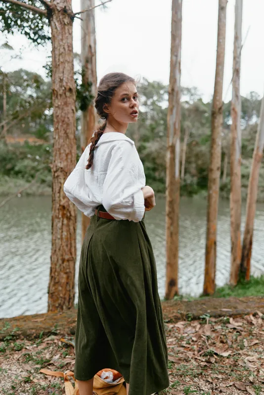 Young woman in a flowing green skirt and white top standing by a tranquil lake surrounded by tall trees, expressing a thoughtful emotion, perfect for seasonal themes