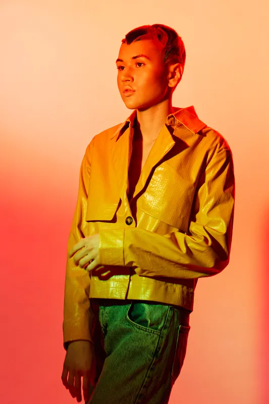 Young woman with short dyed hair wearing a yellow jacket and jeans stands thoughtfully against a warm gradient orange background, showcasing bold contemporary fashion.
