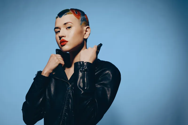 Young woman with colorful painted hairstyle wearing black leather jacket posing confidently against blue background, showcasing bold style and modern fashion attitude.