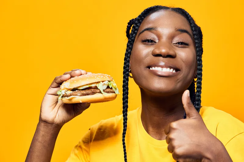 Happy young woman with braids holding a delicious burger, smiling against a vibrant yellow background, showcasing joy in healthy eating, summer vibes