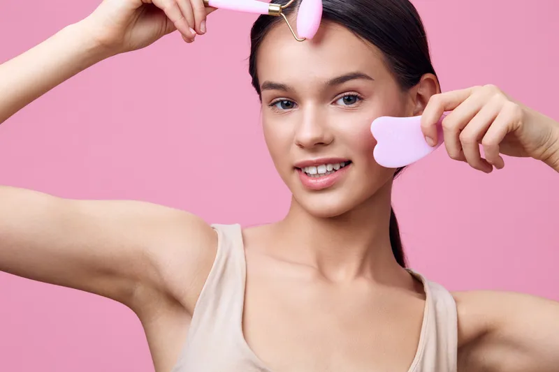 Young woman with a joyful expression holding beauty tools on a soft pink background, focusing on skincare and self care routines