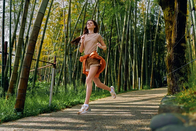 Woman running and jogging on a forest bamboo path outdoors, fitness activity with jacket tied at waist, casual shorts and sneakers, morning nature trail exercise and scenic greenery.