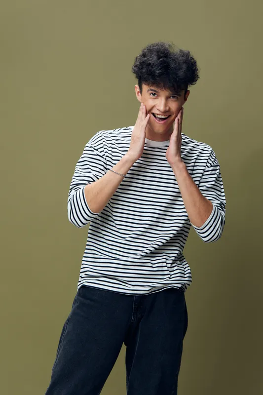 smiling man with curly hair wearing striped shirt and black pants expressing surprise with hands near face on olive background, casual style and youth energy