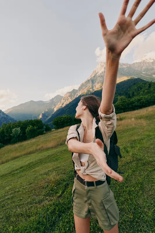Cheerful woman outdoors reaching forward, sunlit moment, relaxed stance, fresh breeze, natural colors, scenic hillside with mountains in the background