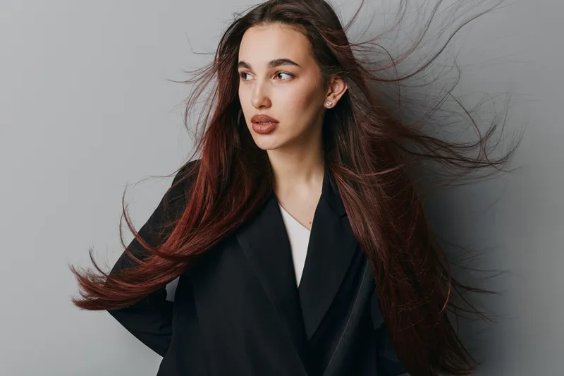 Young woman with long flowing hair and natural makeup posing in black coat against grey background for fashion portrait and beauty concept with confident expression.