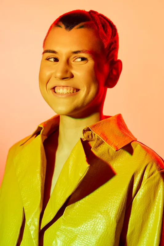 smiling woman with braided red hair wearing a yellow leather jacket, looking sideways with warm orange lighting and soft pink background, vibrant fashion portrait