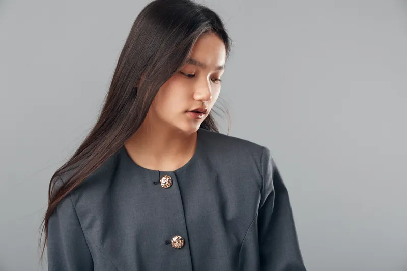 Young woman with long dark hair wearing gray jacket looking down on plain background. Portrait of thoughtful female model with natural makeup and casual style indoors.