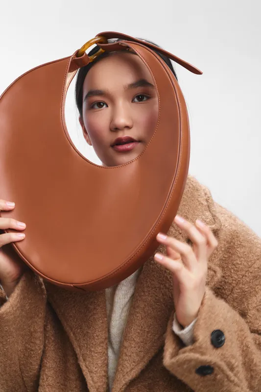 asian woman poses with a brown circular handbag held around her face in a fashion studio, presenting a playful and elegant mood with soft lighting.