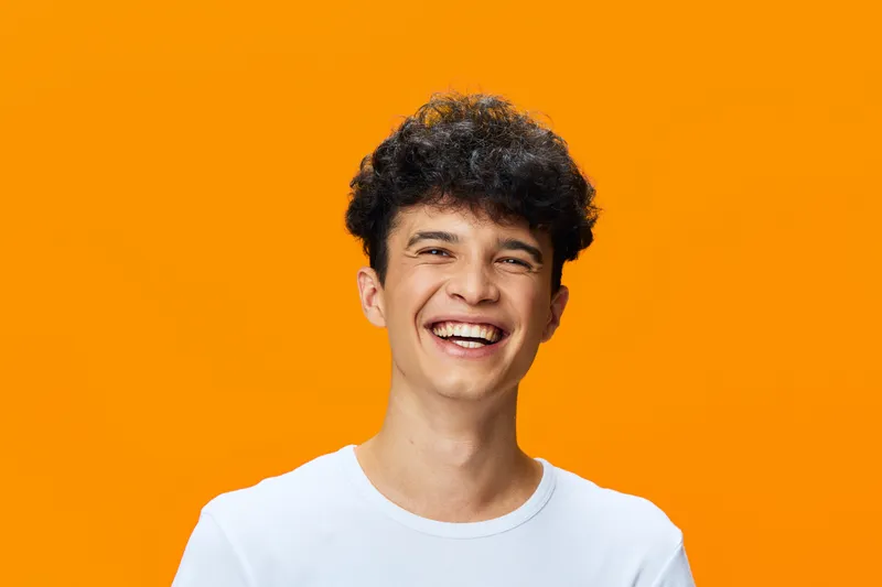 smiling young man with curly hair wearing white shirt on vibrant orange background expressing happiness and joy in casual style