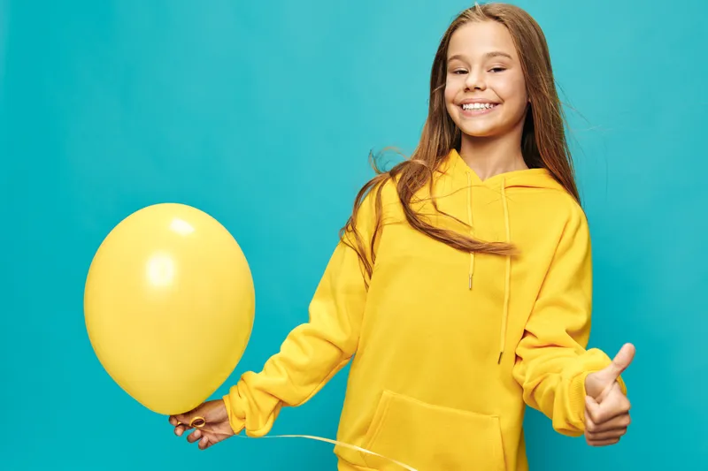 Smiling girl in yellow hoodie holding a balloon and showing thumbs up on turquoise background. Happy child with long hair in casual clothes expressing positivity and joy.