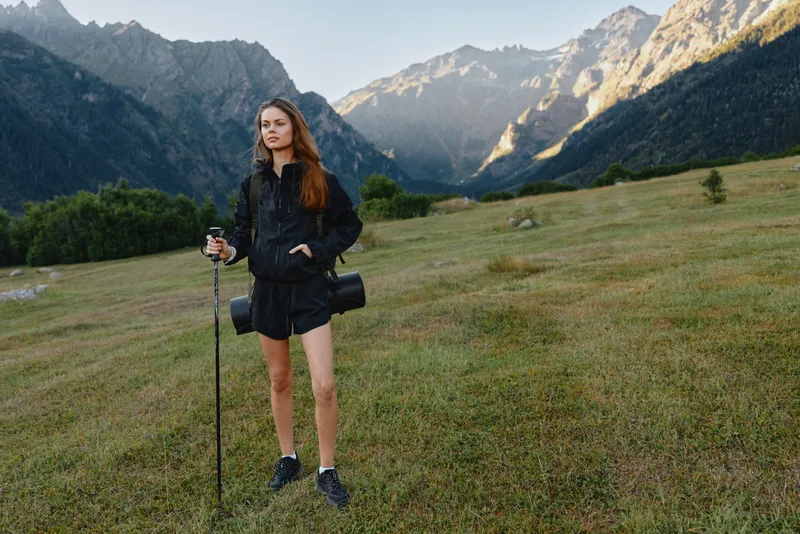 A cheerful young woman stands in a grassy meadow with a hiking pole and backpack, framed by rugged mountains, conveying adventure, focus, and outdoor freedom.