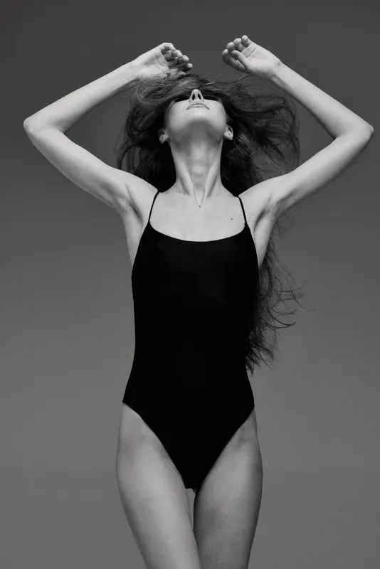 Woman posing in black swimsuit with raised arms in black and white portrait, elegant and expressive.