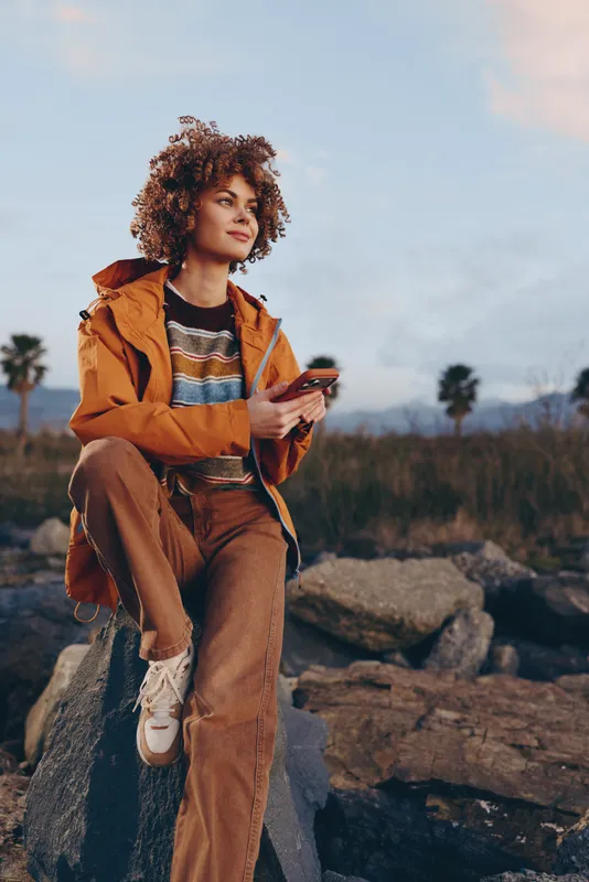 Woman with curly hair outdoors, wearing a casual jacket and holding a phone, standing on rocky landscape at sunset, nature lifestyle mood, soft golden light, relaxed travel moment