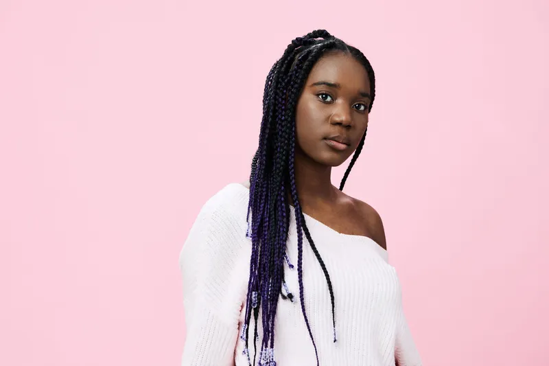 Portrait of a young Black woman with braids, wearing a cozy white sweater against a pink background, expressing confidence and elegance