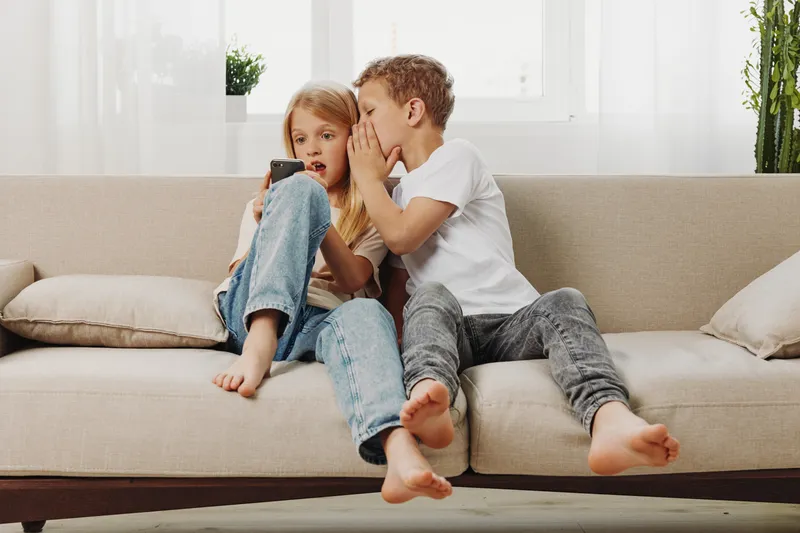 Two children sitting on sofa, boy whispering a secret to girl with surprised expression. Cozy home interior, natural light, concept of friendship and childhood communication.