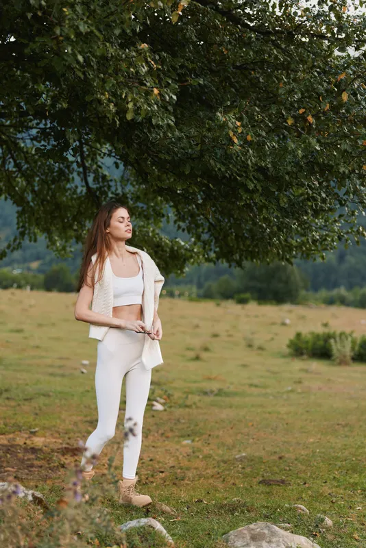 One person, young adult with long hair in white outfit walks through a sunlit field beneath a large tree, a calm, reflective mood in natural light.