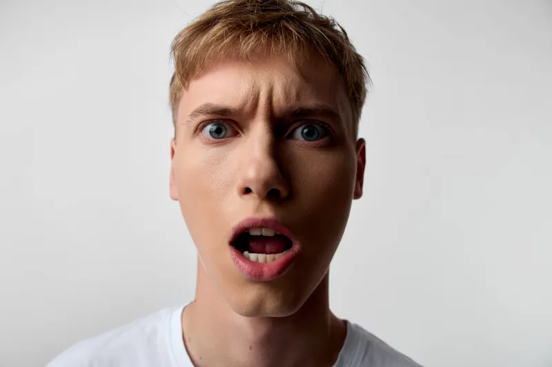 Surprised expression, man, close up, facial reaction, shock, astonishment in a studio shot. Wide eyes, open mouth, strong lighting and neutral background convey sudden reaction and dramatic emotion