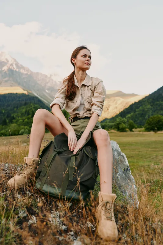 Young woman outdoors sits on a rock in a mountain meadow, wearing hiking boots and a khaki shirt, enjoying a sunlit moment with relaxed confidence in calm light against distant peaks and soft breeze