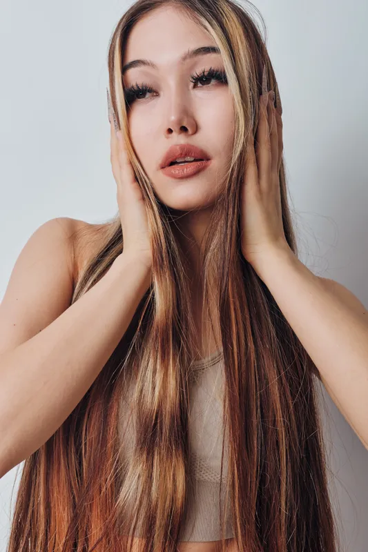 Young woman with long hair and natural makeup posing with hands on face against plain background. Beauty portrait with confident expression and stylish hairstyle indoors.
