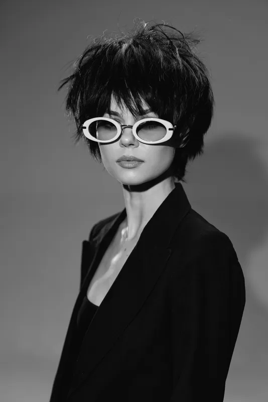 Black and white portrait of a woman with short hair and white sunglasses wearing a dark jacket with a neutral expression