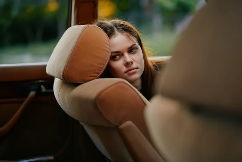 Thoughtful young woman resting her head on the seat in a car, reflecting emotions, surrounded by nature in a warm color palette, capturing a moment of contemplation.