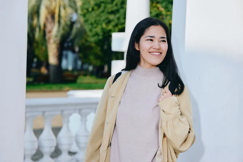 Woman in a beige coat and light sweater stands beside white columns in a sunny outdoor setting, smiling warmly, capturing a casual lifestyle moment suitable for fashion and travel themes