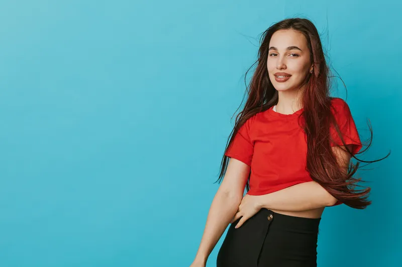 Young woman with long hair wearing red shirt and black pants posing on blue background with confident smile and natural makeup for casual lifestyle portrait photography.
