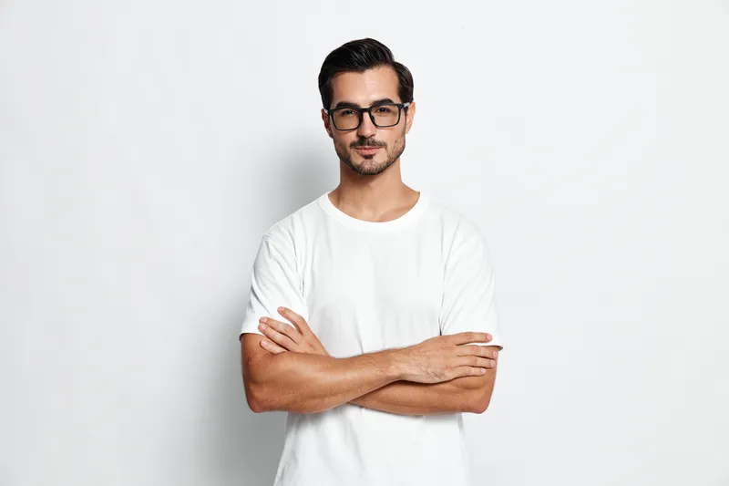 Confident young man stands with crossed arms, wearing a white t-shirt and glasses, looking thoughtfully at the camera against a neutral background.