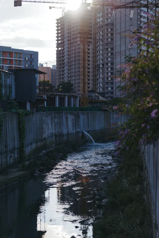 Urban canal with concrete canal and highrise buildings framed by a construction crane at sunset reflection, water stream from a drainage pipe and riverside vegetation in moody twilight.