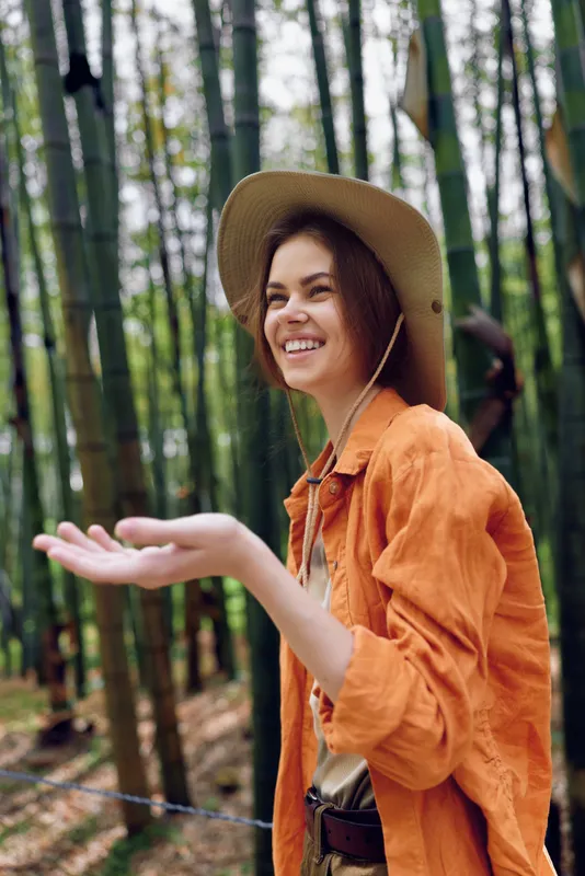 Woman with hat and smile in a bamboo forest, nature travel portrait during hiking trip, wearing orange jacket and enjoying outdoors adventure and eco exploration.