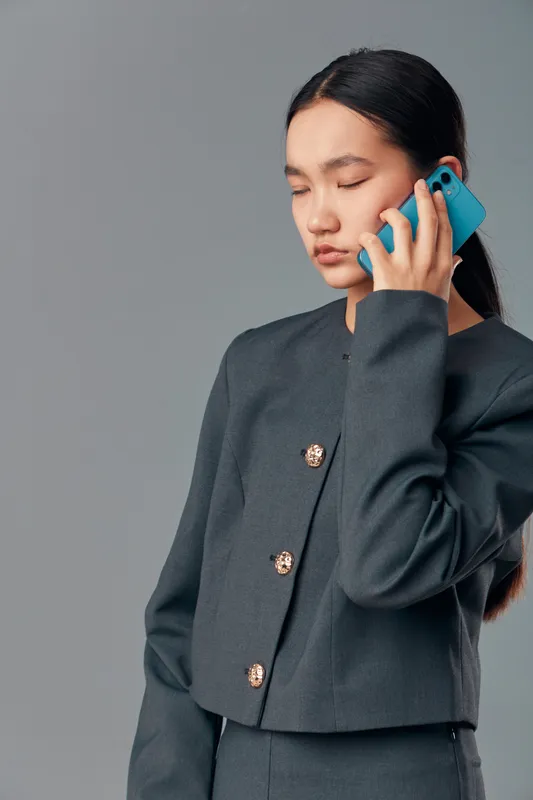 Young woman with long dark hair wearing dark gray suit talking on blue smartphone with eyes closed isolated on gray background for communication technology concept.