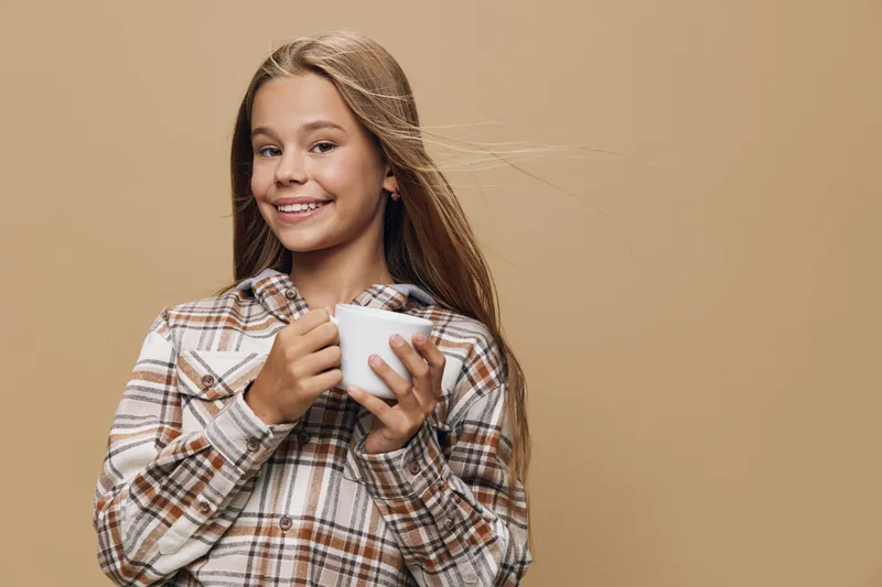 Teenage girl with long hair smiling and holding a white cup wearing a plaid shirt on beige background, cheerful young female enjoying a warm beverage indoors.