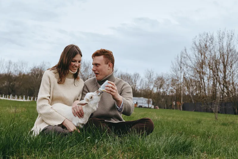 A couple sits on green park grass in cozy sweaters, sharing a playful moment with a fluffy dog, enjoying a relaxed outdoor afternoon together in natural light