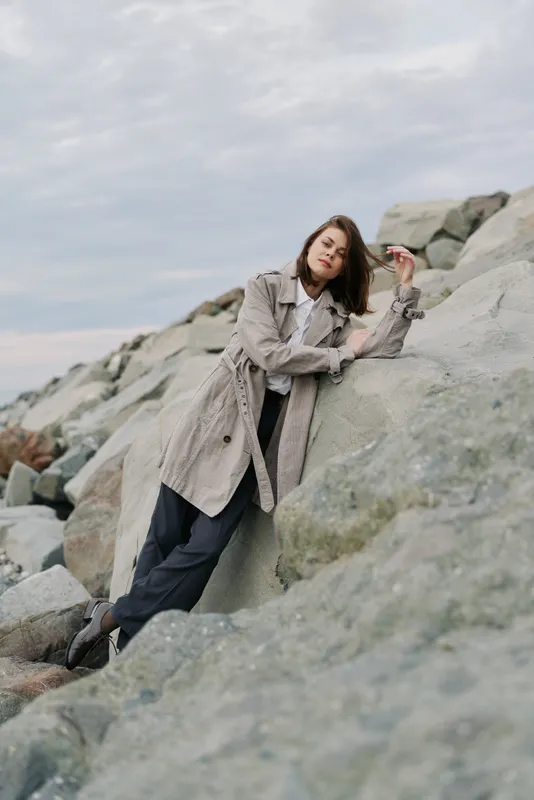 Young woman in trench coat leans on large rocks outdoors. Casual fashion style with relaxed pose on a cloudy day, natural background with stones. Concept of leisure and nature.