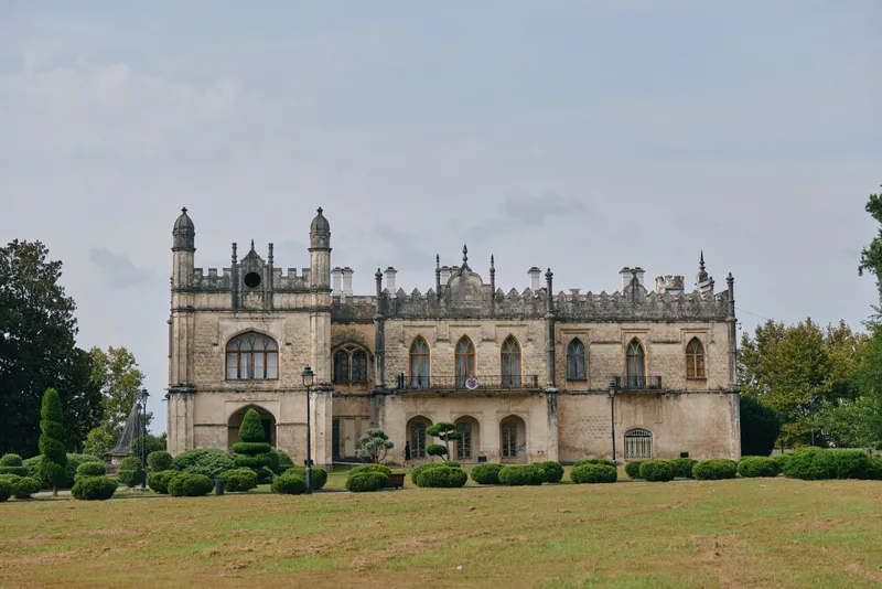Castle building with ornate Gothic facade sits on a green lawn under a blue sky, showcasing historic architecture and tranquil landscape suitable for tourism.