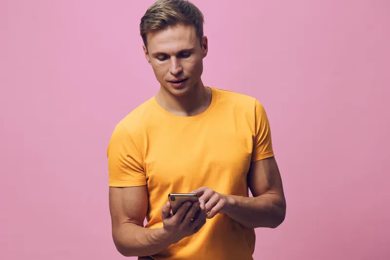 Smiling man with short hair wearing yellow t-shirt using mobile phone isolated on pink background. Casual lifestyle concept, young adult, communication, technology.