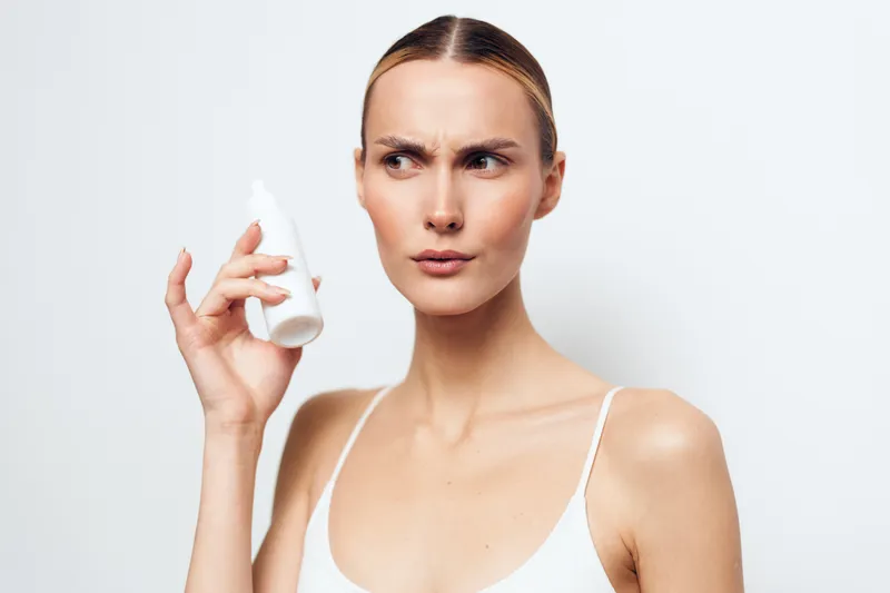 Young woman with clear skin holds white cosmetic bottle near face looks confused or skeptical isolated on white background wearing white tank top natural beauty concept.