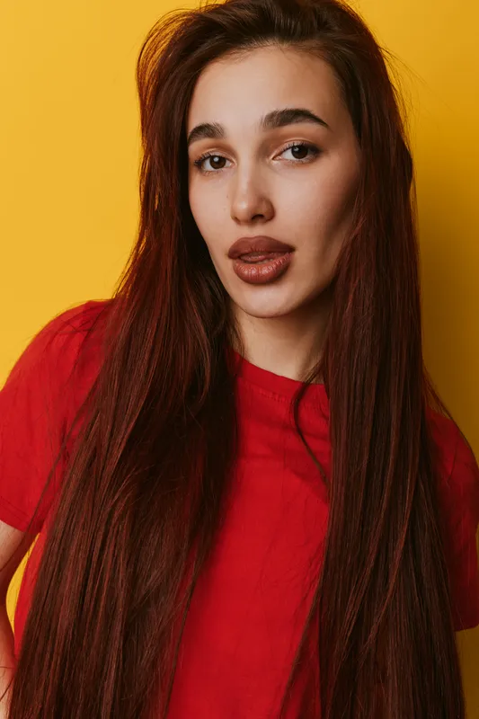 Young woman with long brown hair wearing red shirt posing against yellow background. Portrait of female model with natural makeup and serious expression in studio lighting.