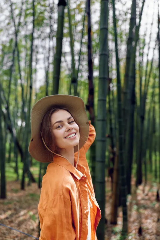 Woman smile in a bamboo forest with hat and orange jacket, portrait outdoors in nature. Joyful traveler posing among tall green bamboo stalks with soft summer light and relaxed expression.