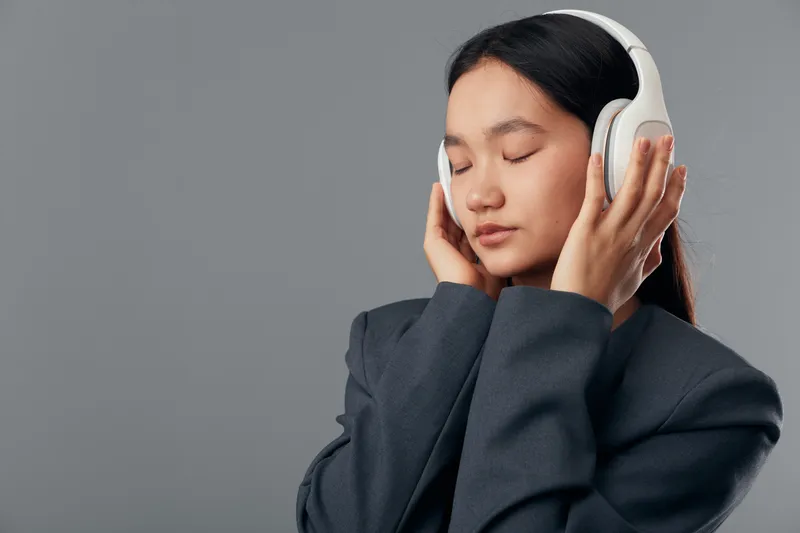 Young woman wearing white headphones with closed eyes, enjoying music in gray jacket. Portrait with calm expression on plain gray background for relaxation and sound experience.
