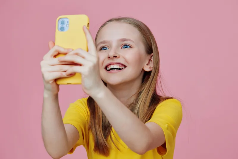 Happy girl taking a selfie with a yellow phone on a pink background, embodying joy and youthfulness in a vibrant setting