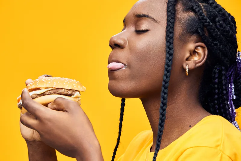 A joyful young African woman savoring a delicious burger against a vibrant yellow background, showcasing happiness and indulgence in fast food