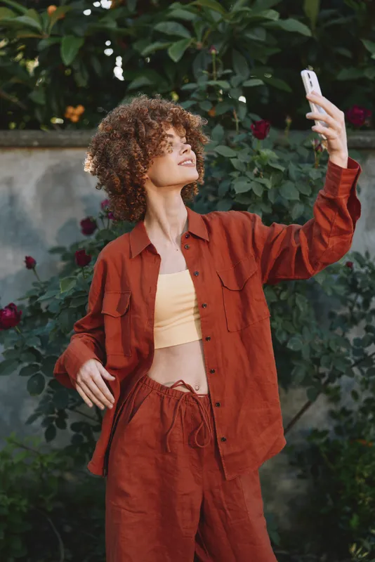 A stylish woman with curly hair takes a selfie in a sunlit garden, wearing rust colored casualwear. The candid moment conveys confidence, self expression and everyday lifestyle