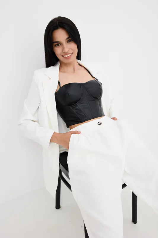 Smiling woman in white pants and blazer with black corset top posing confidently against a plain light background, showcasing modern fashion and elegance.