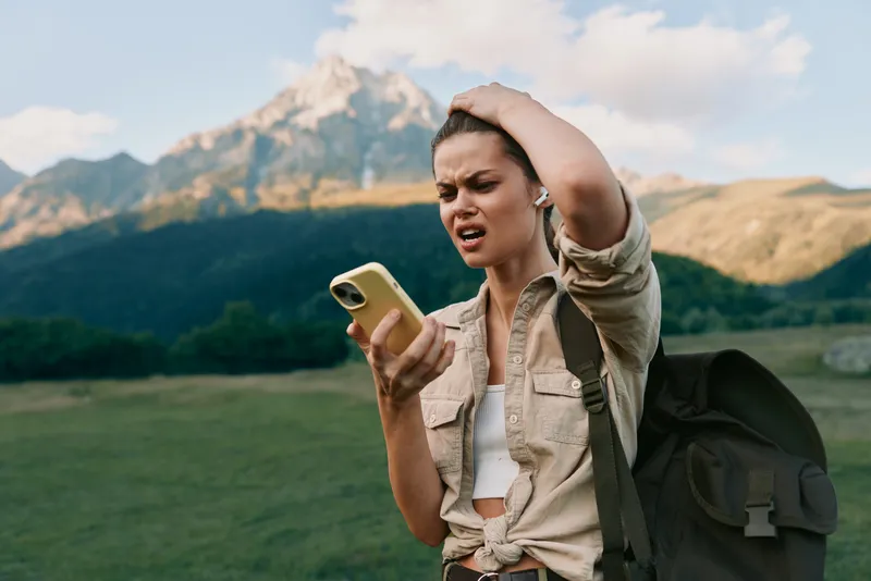 A puzzled woman outdoors studies her phone while gripping her head, backpack slung over one shoulder. A scenic mountain landscape and bright sky set a reflective travel moment.